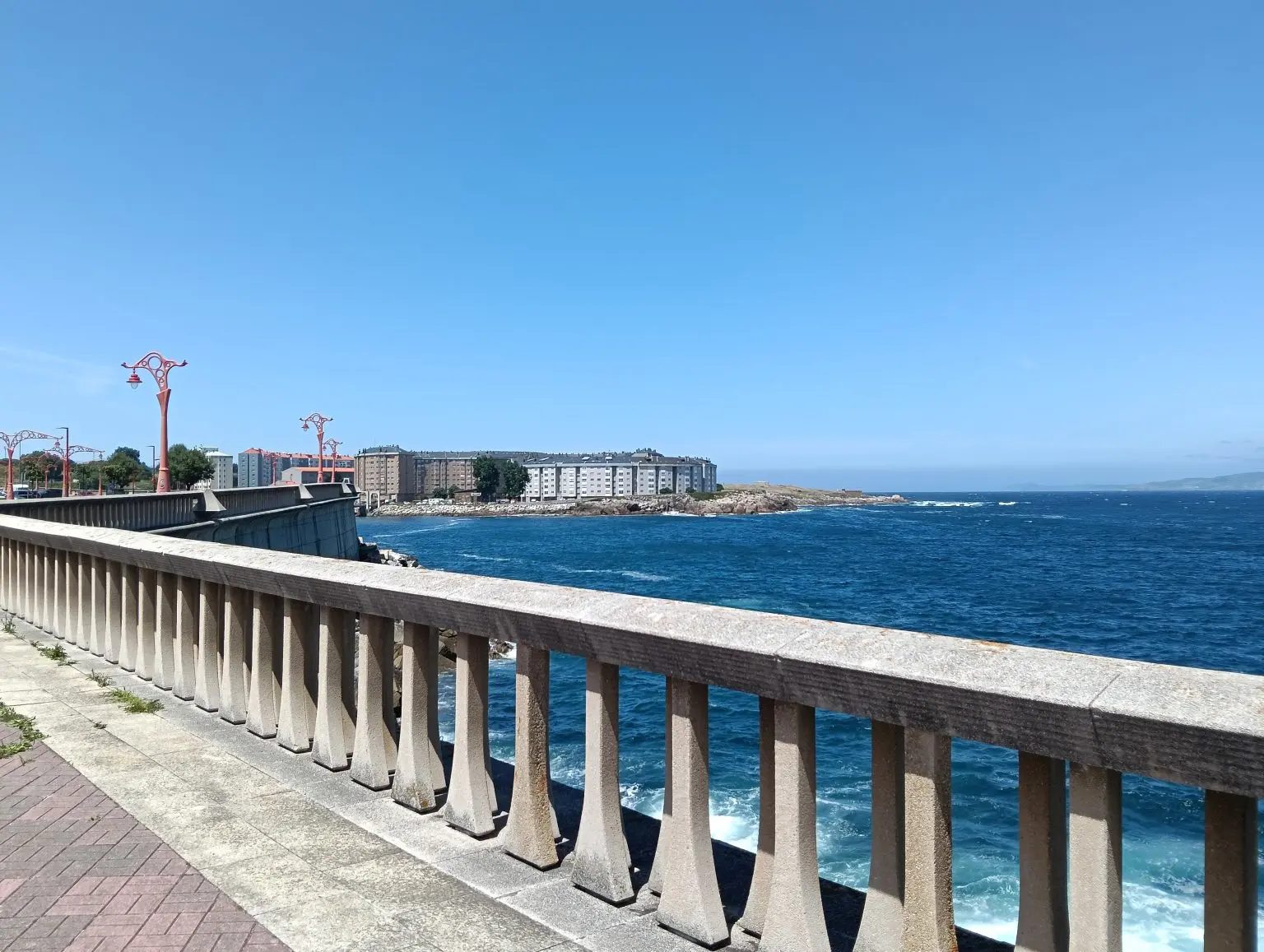 Buildings above the coast and the open sea, seen from a street with a stone railing on a clear day.