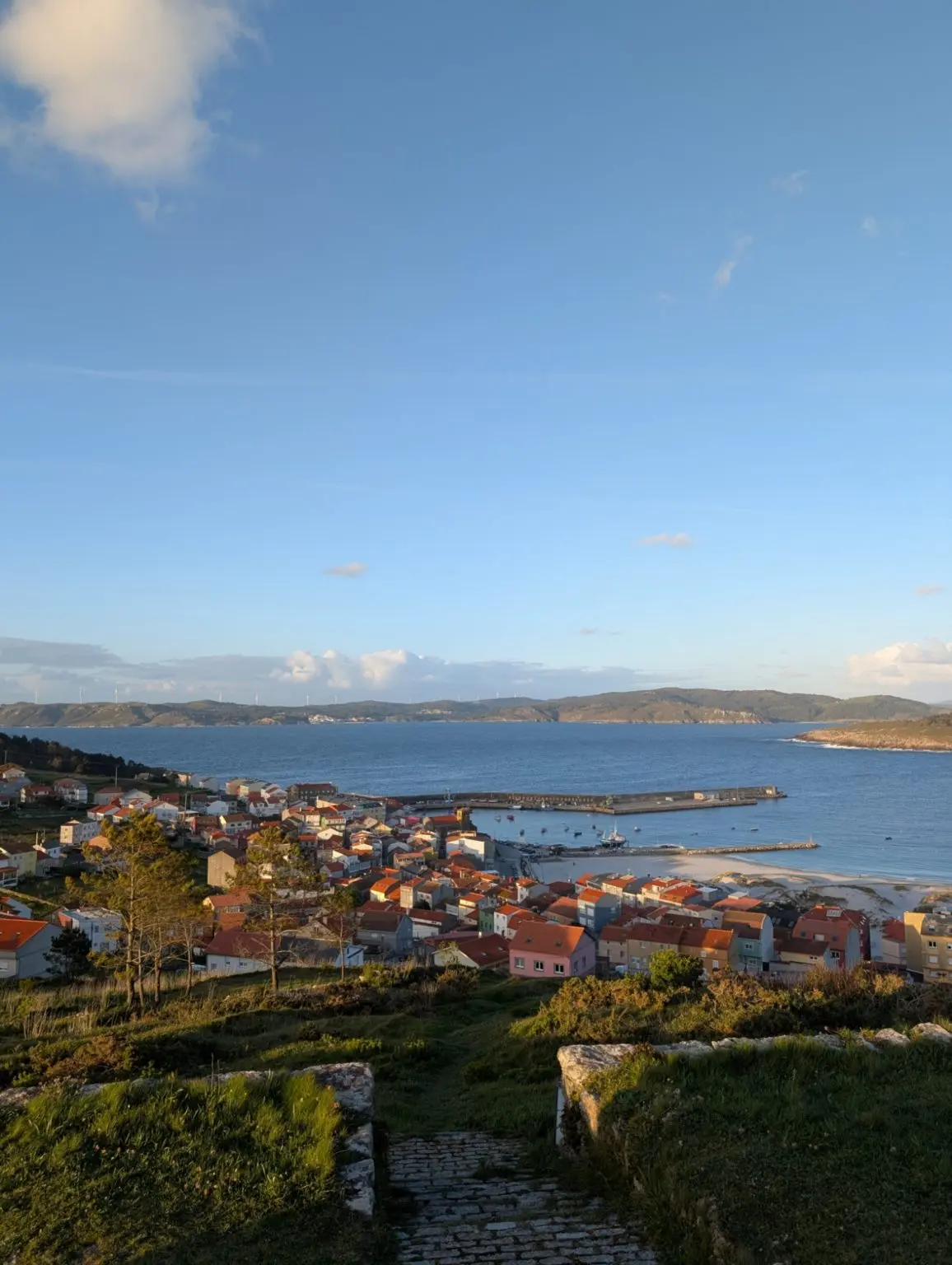 A coastal village of red-roofed houses around a harbour and beach on a blue bay, under a clear afternoon sky.