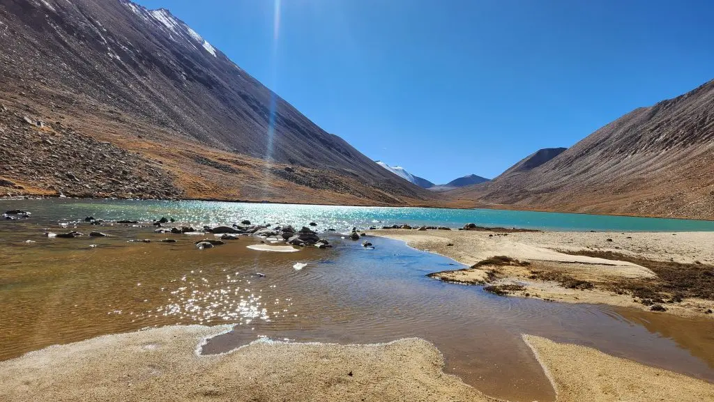 Turquoise lake surrounded by rocky, snow-capped mountains in Langtang, with a sandy shoreline under a clear blue sky.